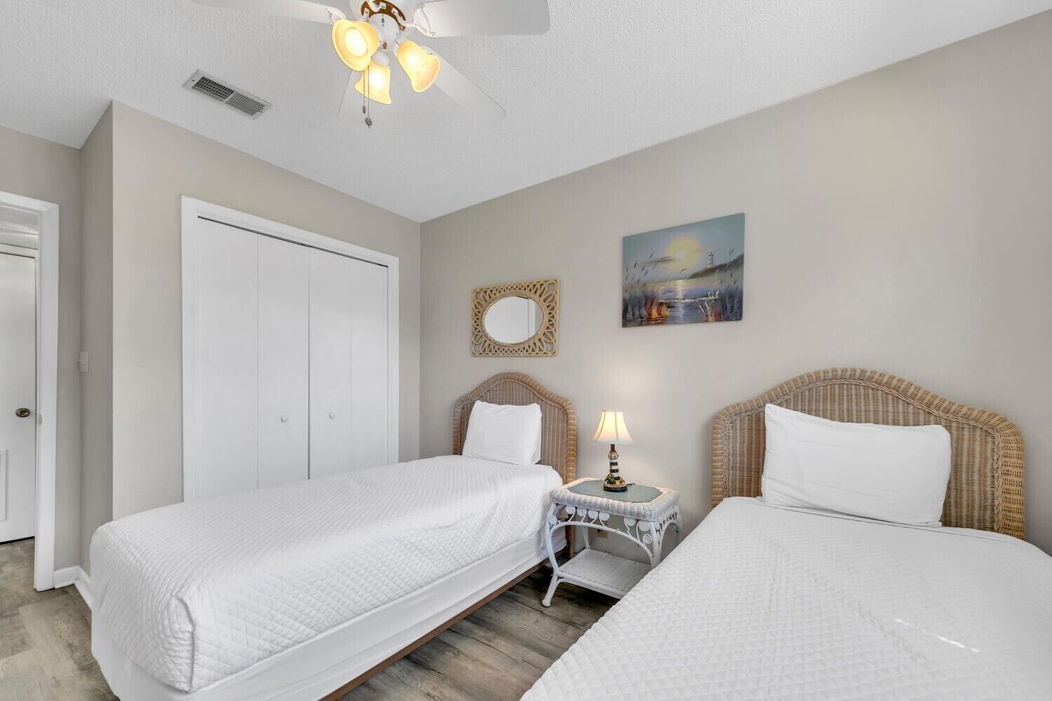 A bedroom with two single beds, a ceiling fan, a small table with a lamp, a door, and a painting on the wall.