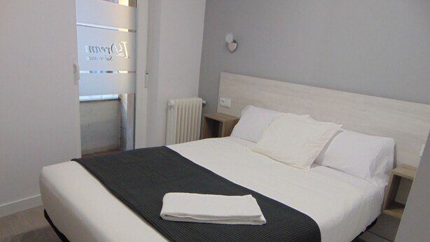 A neatly made bed with white linens and a black throw blanket, a bedside table with a lamp, and a radiator on the wall. - Hostal I Dream Salamanca (Salamanca)