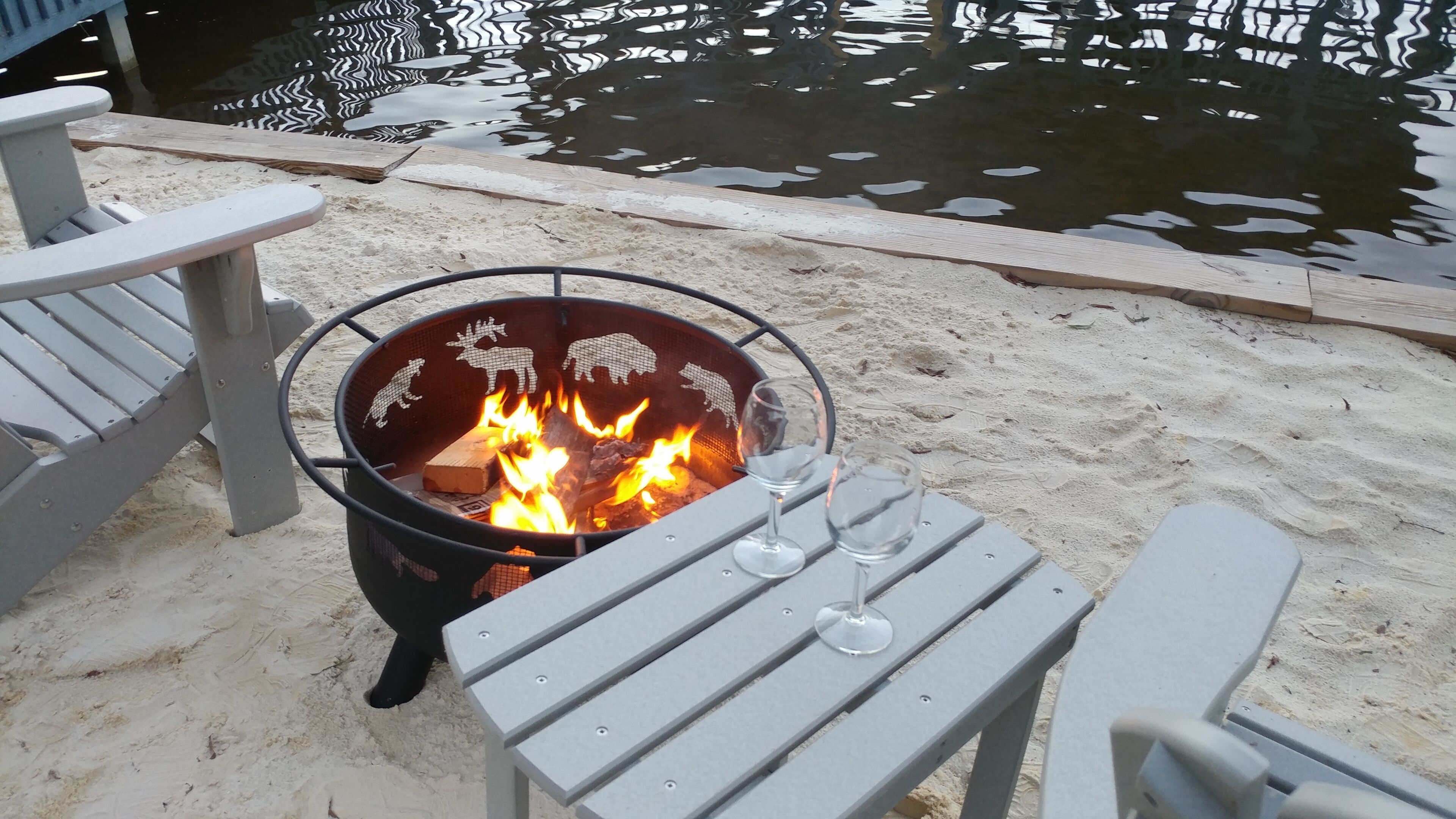 A fire pit with a metal grill and a table with two glasses on it, set on a sandy area near a body of water.
