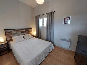 A bedroom with a wooden headboard, a large bed with white linens, a bedside table with a lamp, a window with curtains, and a radiator. - BRIGHT COTTAGE IN THE HEART OF LUBERON. Pool unavailable from July 4 to Sept 1. (Forcalquier)