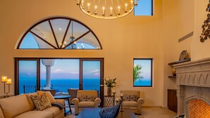 Ein geräumiges Wohnzimmer mit großem Bogenfenster mit Meerblick, Kamin und einer Kristallleuchter.