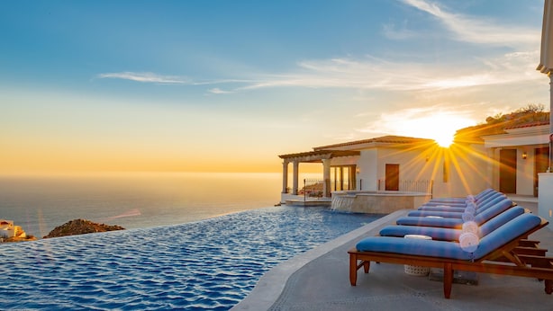 Ein Infinity-Pool mit Meerblick und Sonnenuntergang, Liegestühle und ein überdachter Bereich mit Säulen.