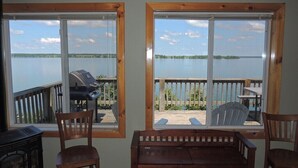 A room with two large windows overlooking a lake, a wooden deck, and a barbecue grill outside.