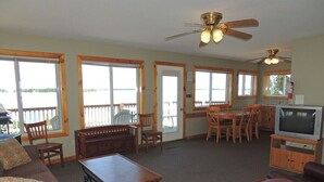 A spacious living room with a sofa, a leather chair, a wooden coffee table, and a television on a wooden stand. There are multiple windows providing views of the outdoors.