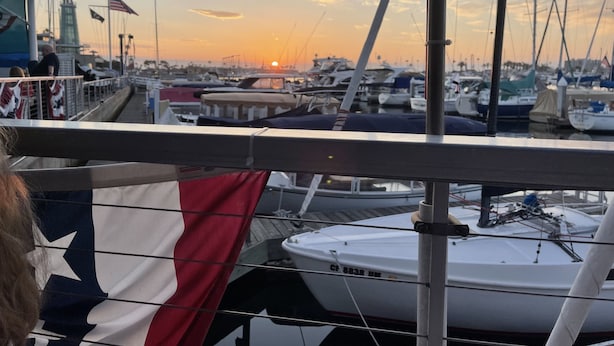 Uma marina com vários barcos atracados, uma bandeira e um farol ao fundo.