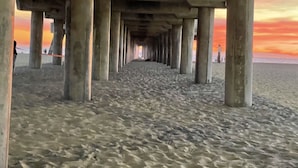 Um cais com pilares de concreto que se estende até a areia, dando acesso a uma praia onde se veem pessoas ao longe.