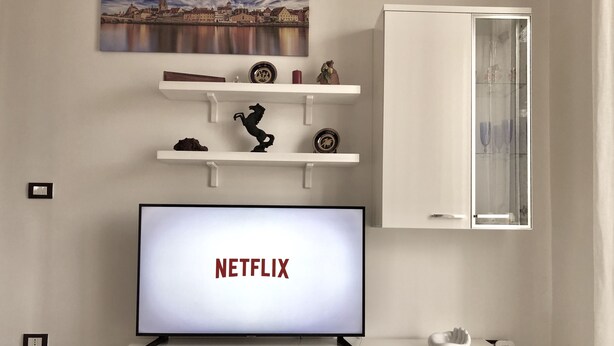 Un salón moderno con un televisor de pantalla plana mostrando "NETFLIX", un armario blanco con puertas de cristal y objetos decorativos en estantes.