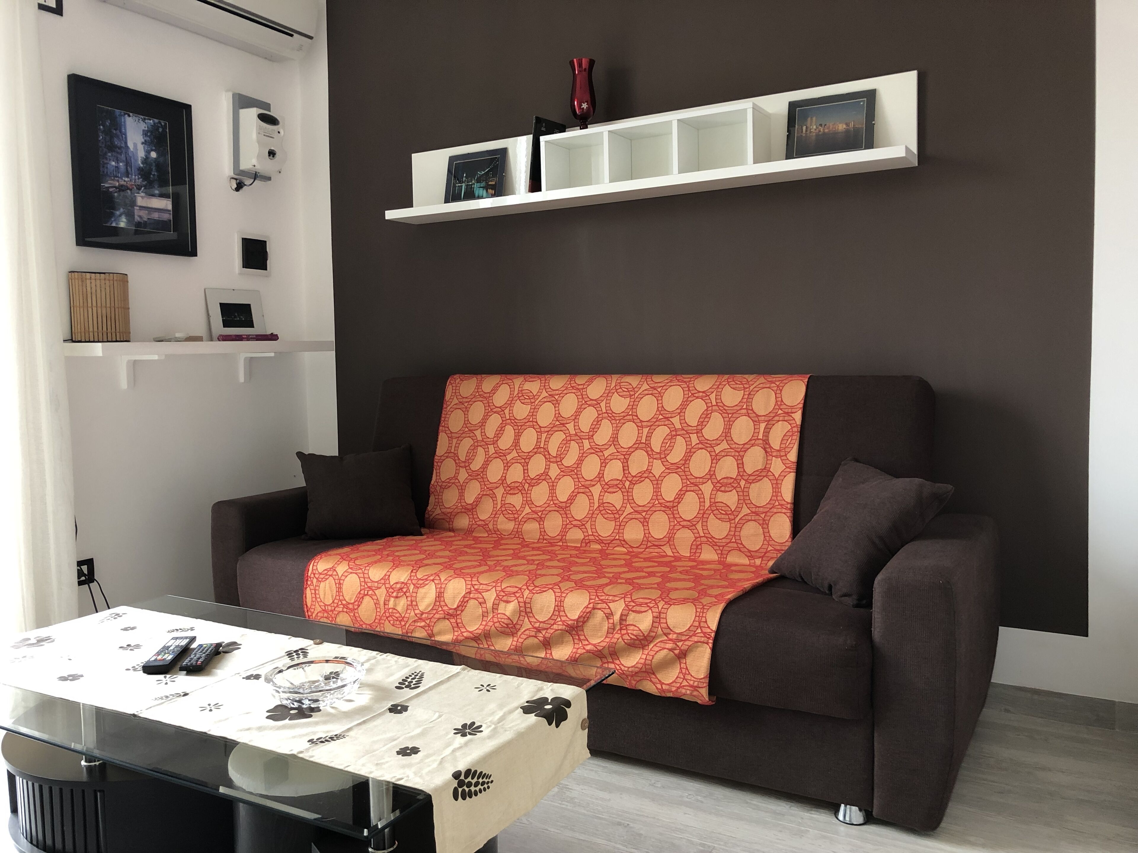 A modern living room with a dark brown sofa, a patterned throw blanket, and a glass coffee table with remote controls and a bowl.