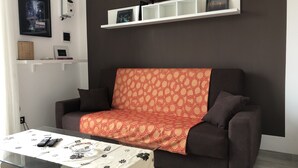 Una sala de estar moderna con un sofá marrón oscuro, un plaid estampado y una mesa de centro de vidrio con controles remotos y un bol.