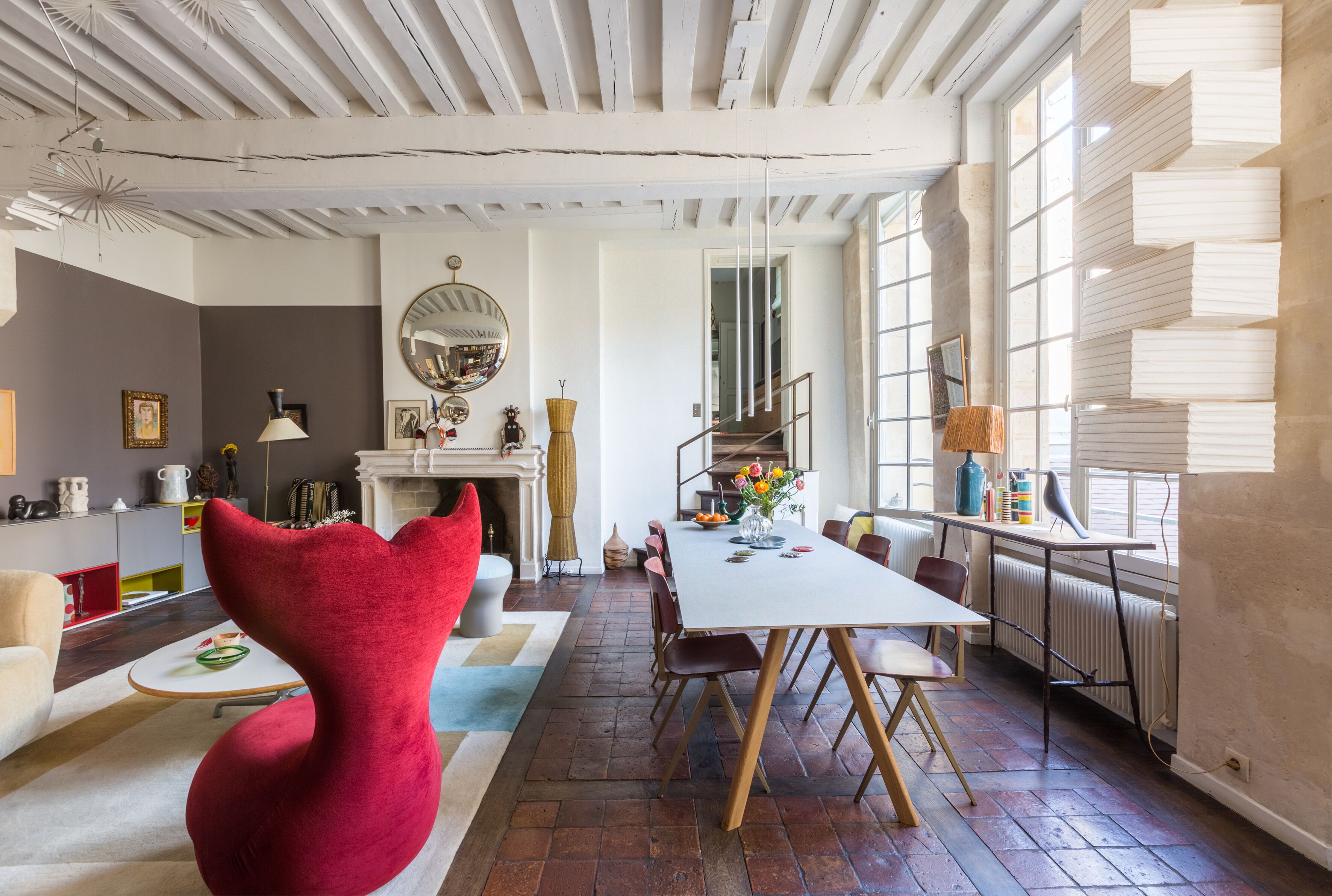 A dining area with a large table, chairs, and a red chair. There is a fireplace, a mirror, and a window with blinds.