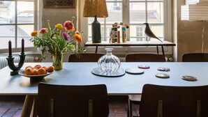 A dining area with a table set for two, a vase with flowers, and a view of buildings through large windows. - Beautiful Loft on Rue Quincampoix, by Veeve (Paris)