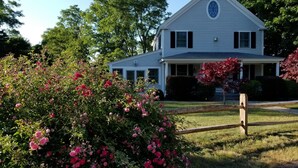 Une maison à deux étages avec un porche, entourée d’une végétation luxuriante et d’arbustes en fleurs.