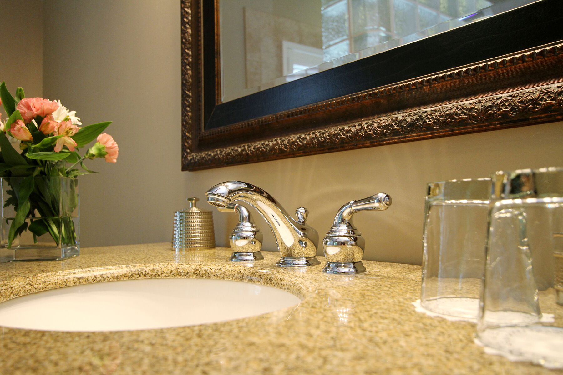 A bathroom with a granite countertop, a sink, two faucets, a mirror with a wooden frame, and a vase with flowers.