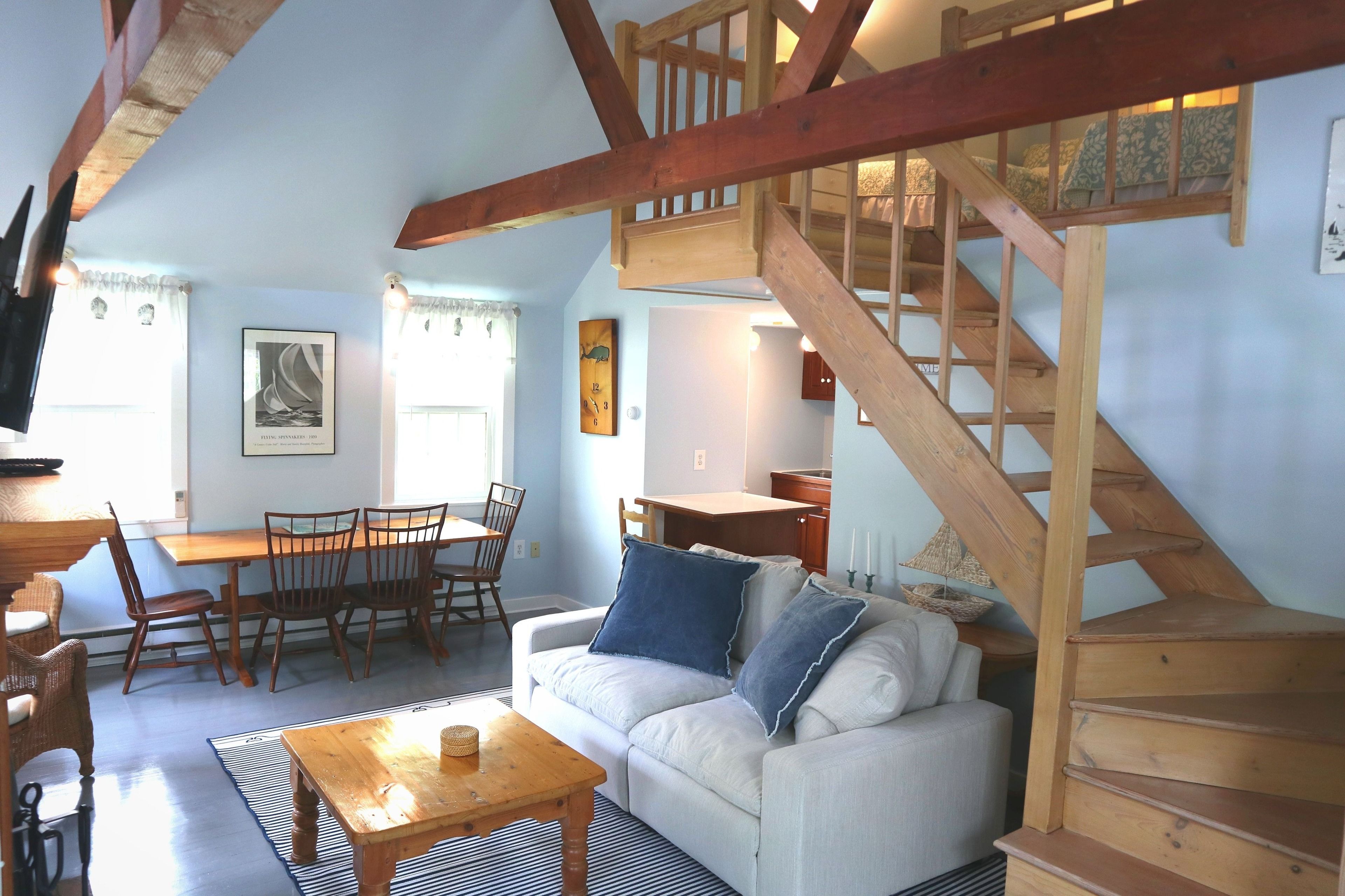 A cozy living room with a wooden staircase, a dining area with chairs and a table, and a sofa with cushions.