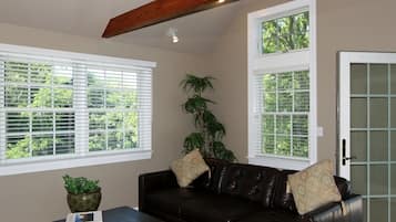 A living room with a dark leather sofa, two windows with blinds, a potted plant, and a door with frosted glass panels.