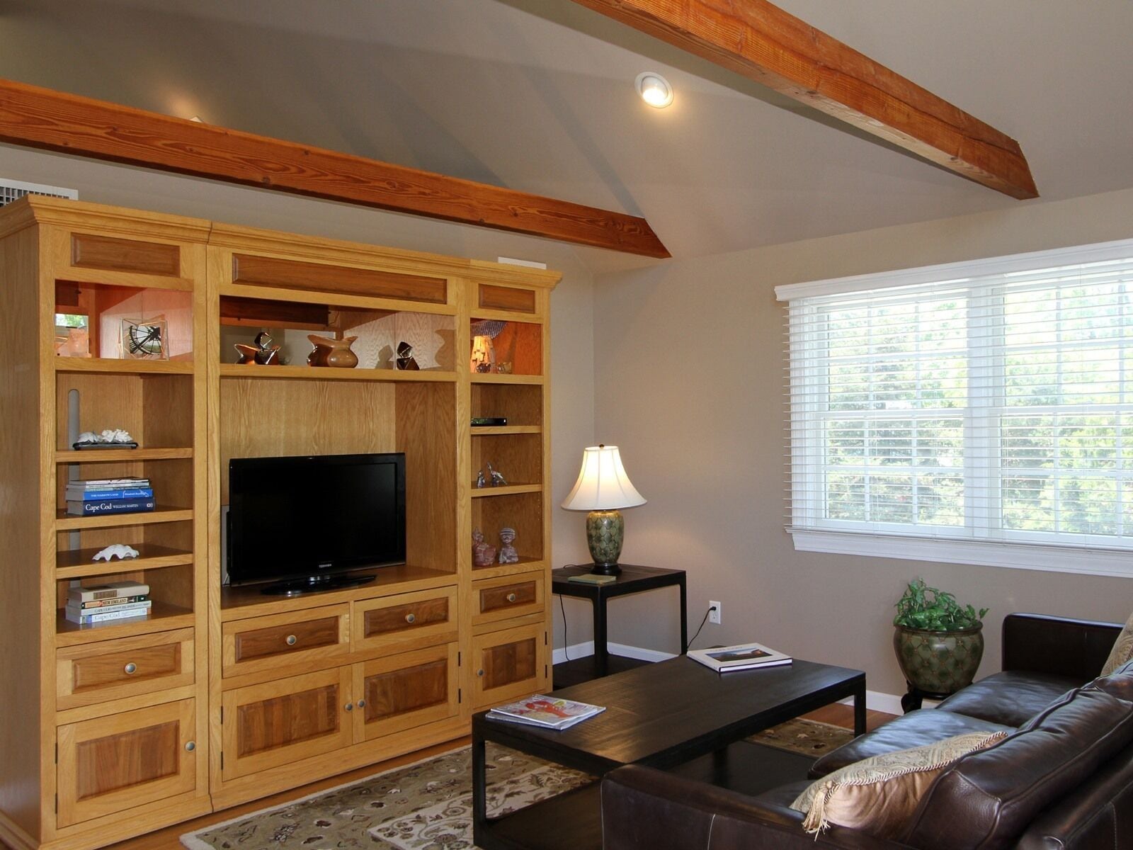Una sala de estar con un mueble de madera para entretenimiento, un televisor de pantalla plana, una mesa de centro, un sofá y una ventana con persianas.