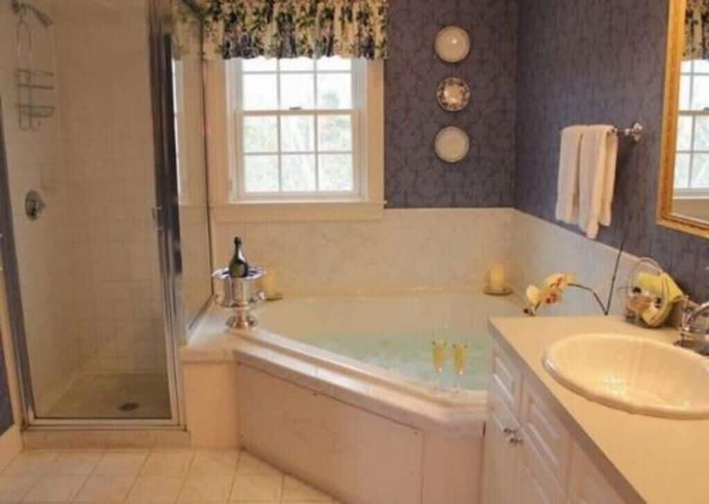 A bathroom with a glass-enclosed shower, a bathtub with two faucets, a sink, and a window with a view of trees.