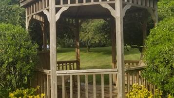 A wooden gazebo with a covered porch and a small table, surrounded by greenery and flowers.