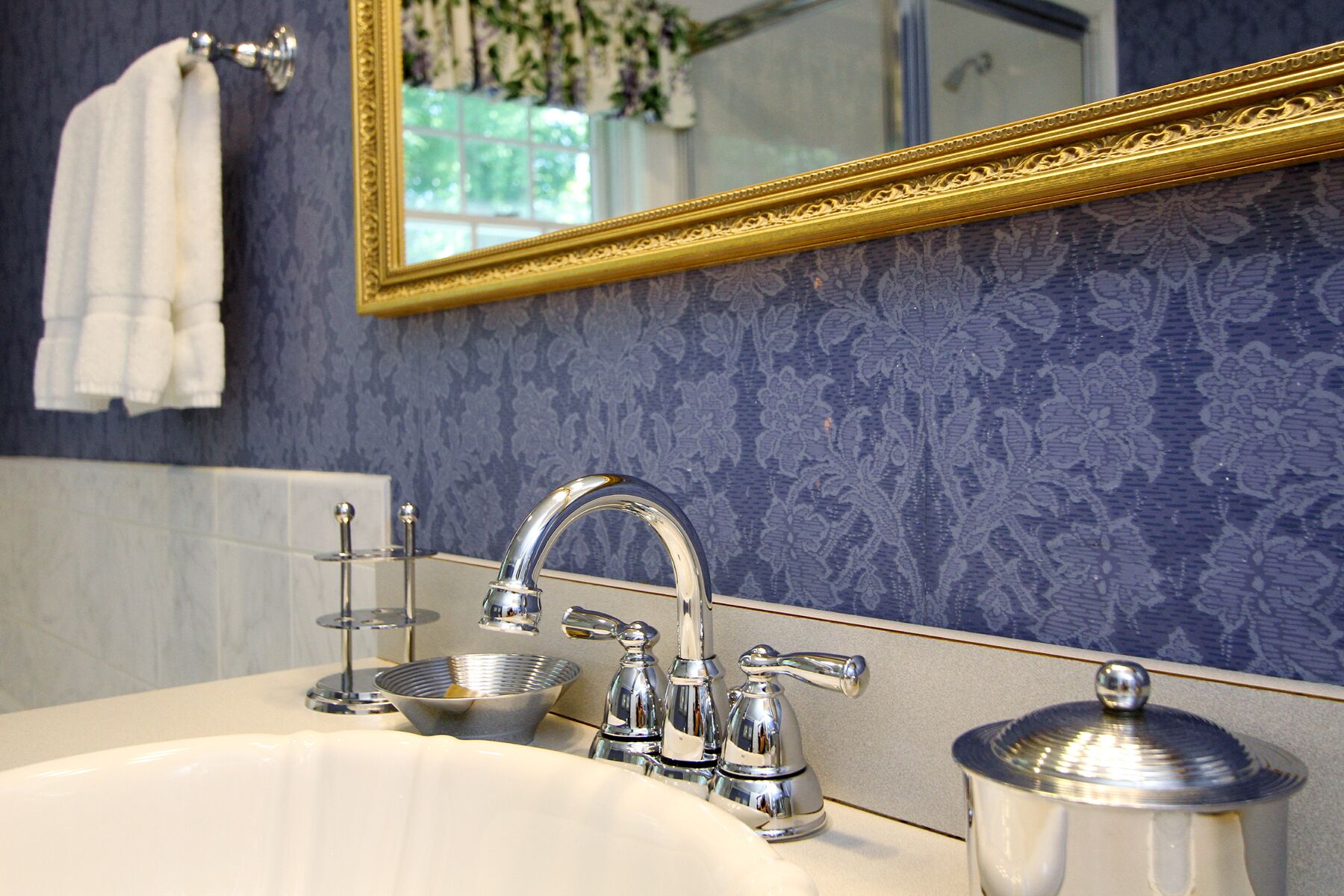 Une salle de bain avec un lavabo blanc, des robinets chromés, un miroir à cadre doré et un mur orné d’un motif bleu.