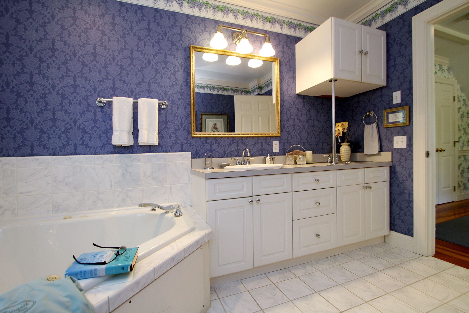 Un baño con bañera, lavabo y tocador con cajones. Hay un espejo sobre el lavabo y una puerta que da a otra habitación.