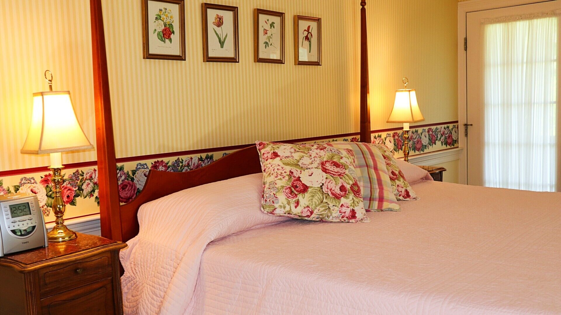 A neatly made bed with floral pillows, a bedside table with a clock, and framed botanical prints on the wall.