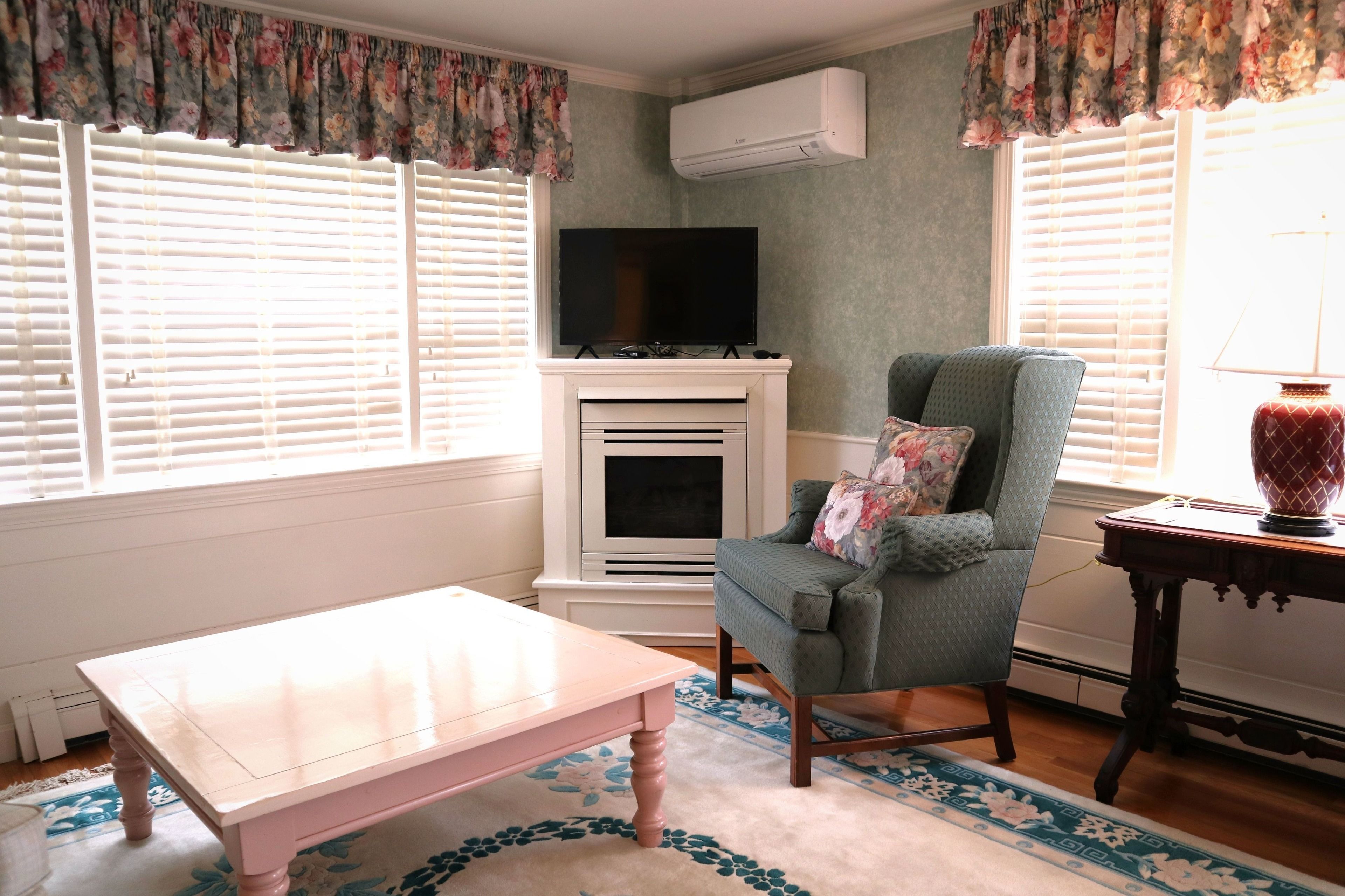 Un salon avec un téléviseur posé sur un meuble, un fauteuil vert agrémenté d’un coussin à motifs floraux, une petite table et une lampe sur une table d’appoint.