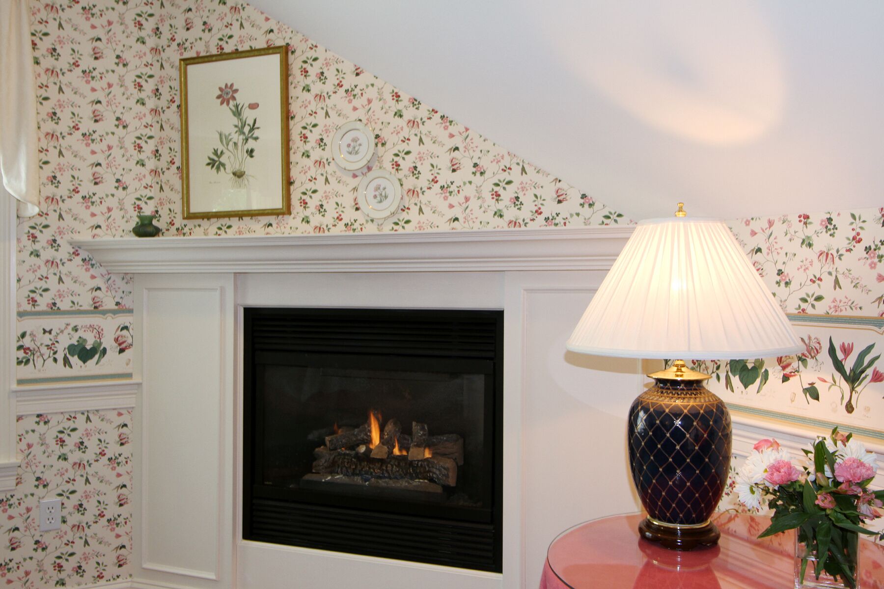 A fireplace with a lit fire, a decorative lamp, and floral wallpaper.