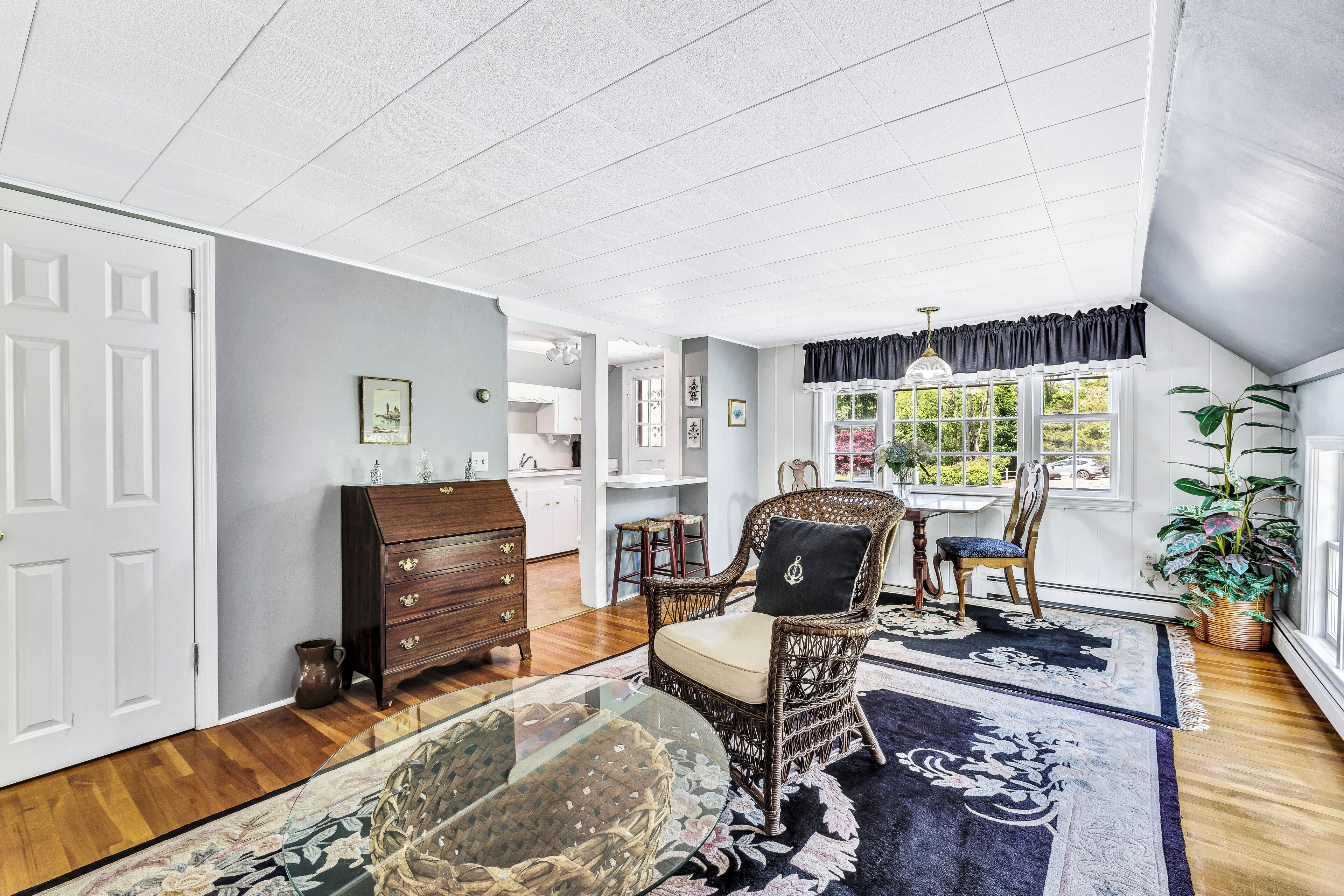 A spacious room with a large glass table, wicker chairs, a dark blue rug, and a view of the outdoors through a window.