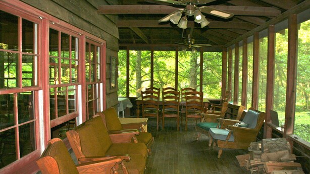 A covered porch with wooden furniture, a ceiling fan, and large windows offering a view of the surrounding forest. - Large SECLUDED RIVERFRONT Cabin with PRIVATE access to CANOE/KAYAK Blue River (Depauw)