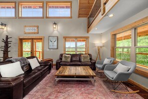 A spacious living room with a brown leather sofa, a wooden coffee table, and grey rocking chairs. There are large windows providing natural light and views of the outdoors. - Custom built home on 5 private acres, unobstructed views, and a hot tub.  (Leavenworth)