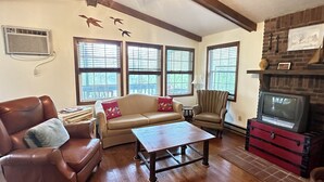 A living room with a brown leather couch, beige sofa, and a wooden coffee table. There are three windows with blinds, a fireplace with a television, and a wall-mounted air conditioner. - Waterfront 2 Bd cabin on Blue River, PRIVATE RIVER ACCESS, kayaking, fishing (Corydon)