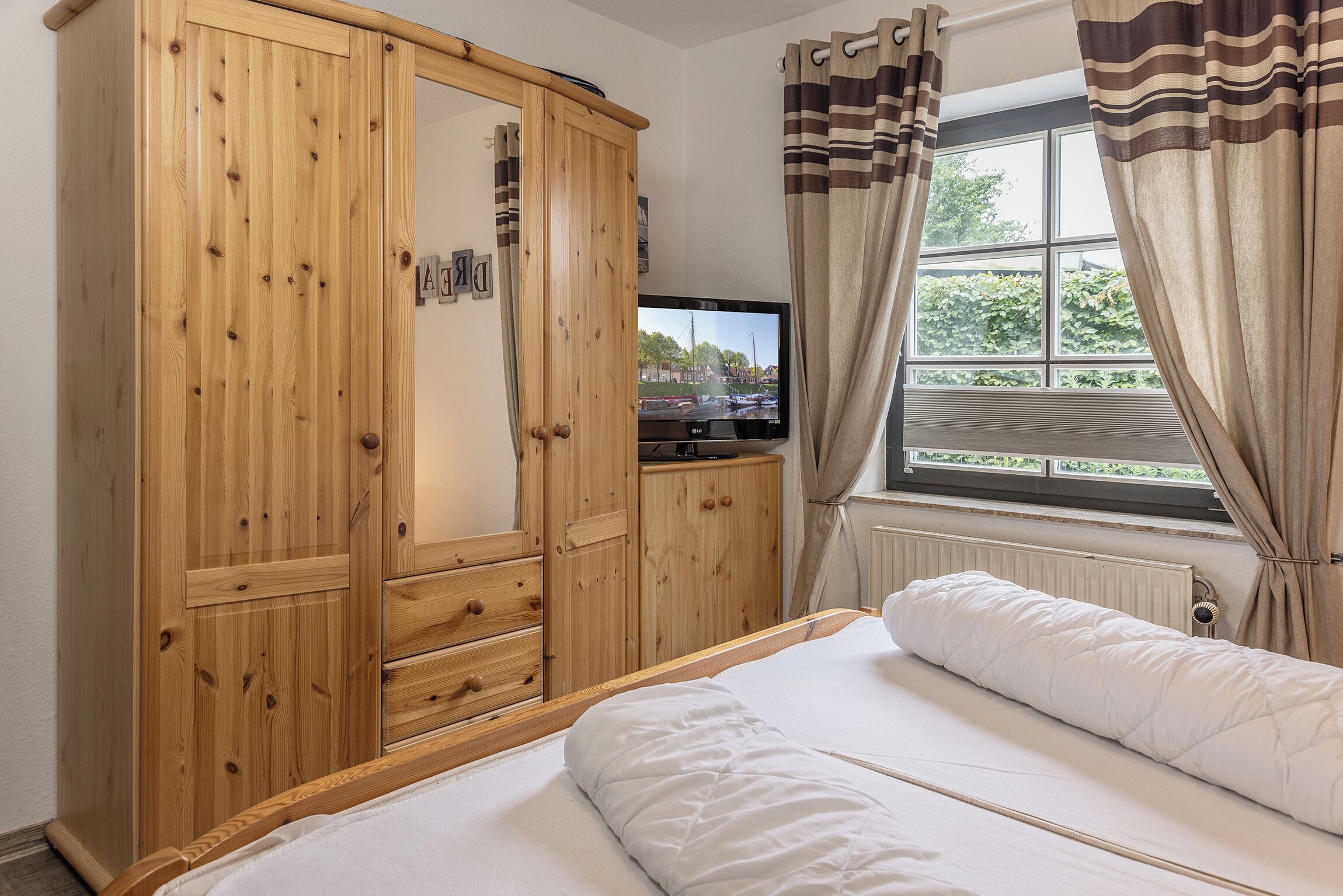 A bedroom with a wooden wardrobe, a television, a bed with white bedding, and a window with curtains.