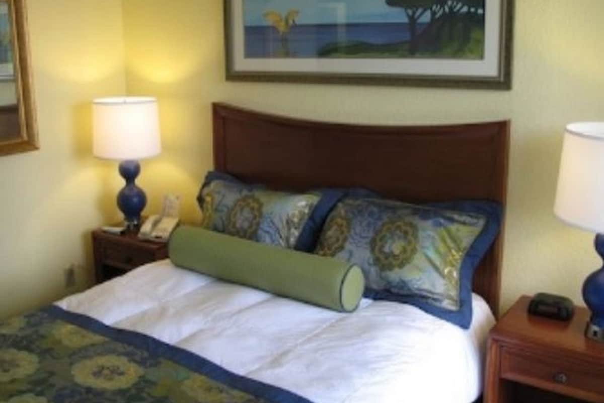 A hotel room with a bed, two bedside tables, a lamp on each table, a painting on the wall, and a window with a view of the sea.