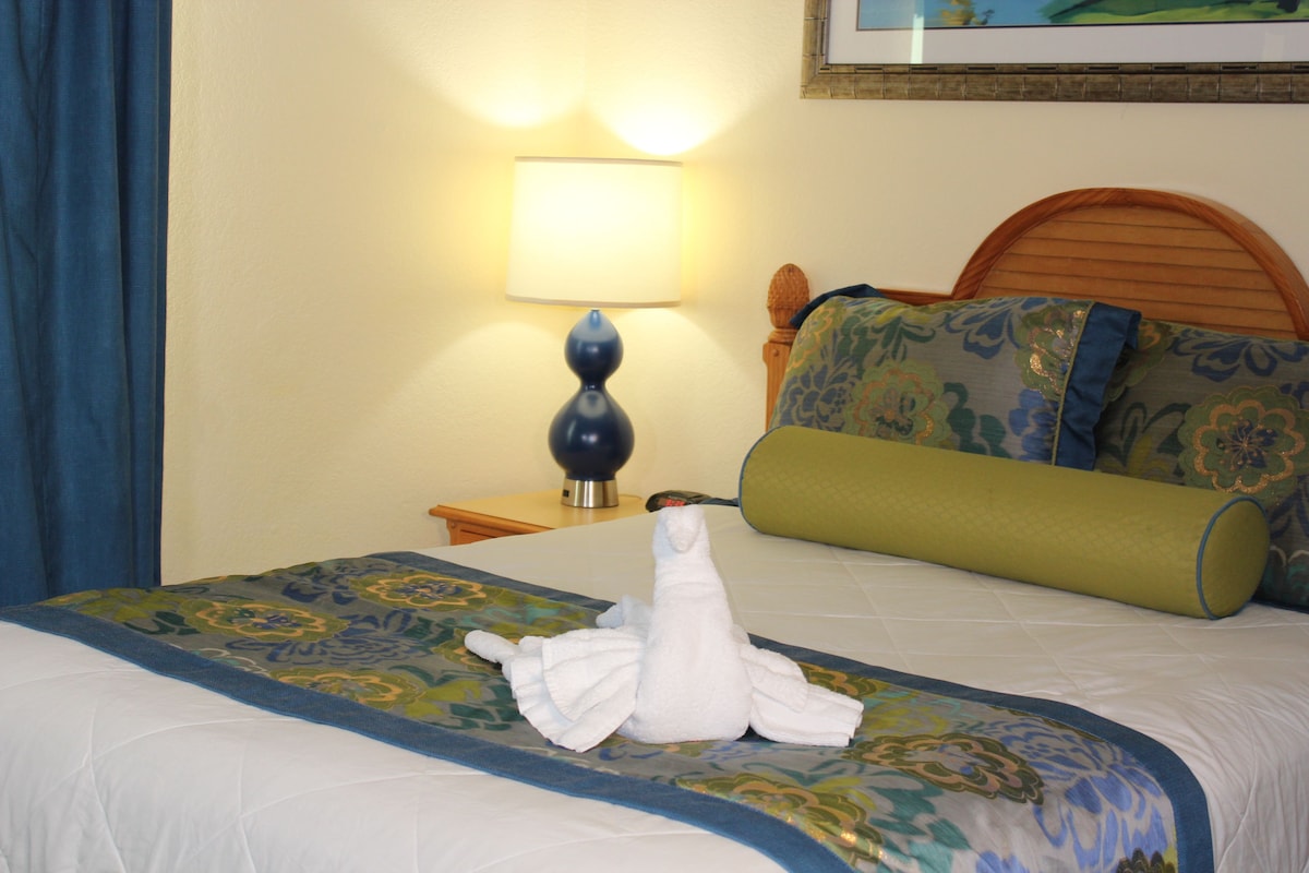 A hotel room with a neatly made bed, a lamp on the nightstand, and a painting on the wall.