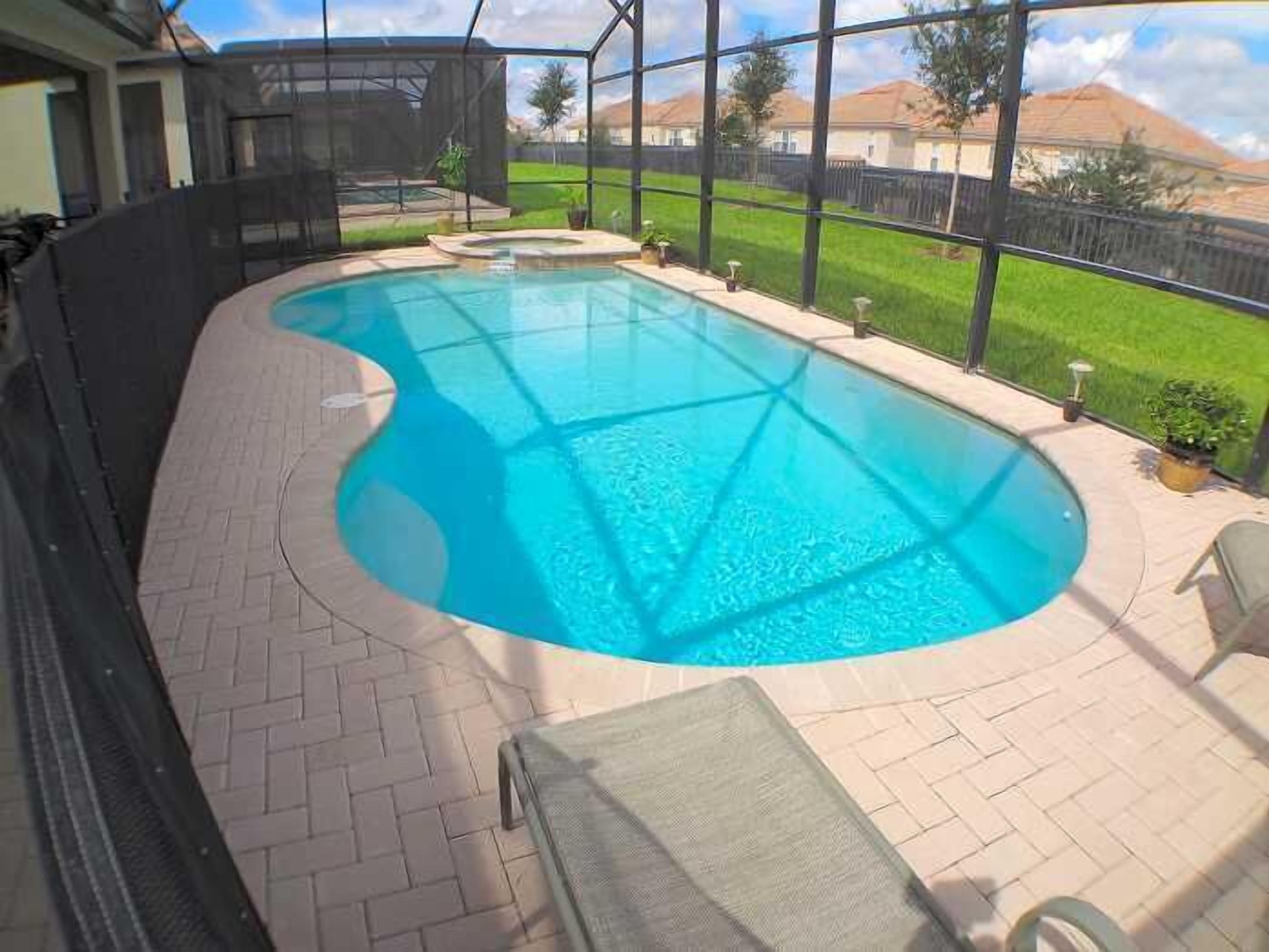 Una piscina con bordo curvo, circondata da un patio in mattoni e un’area coperta con vista su un prato verde e case sullo sfondo.