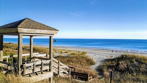 A wooden beach shelter with a roof, located near a beach with clear blue water and a sandy shore.