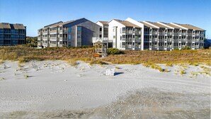 A multi-story building with balconies, situated on a sandy beach with sparse vegetation.