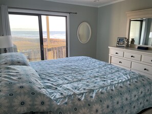 Quarto com cama grande, cômoda, espelho e vista para o oceano através de uma porta de correr.
