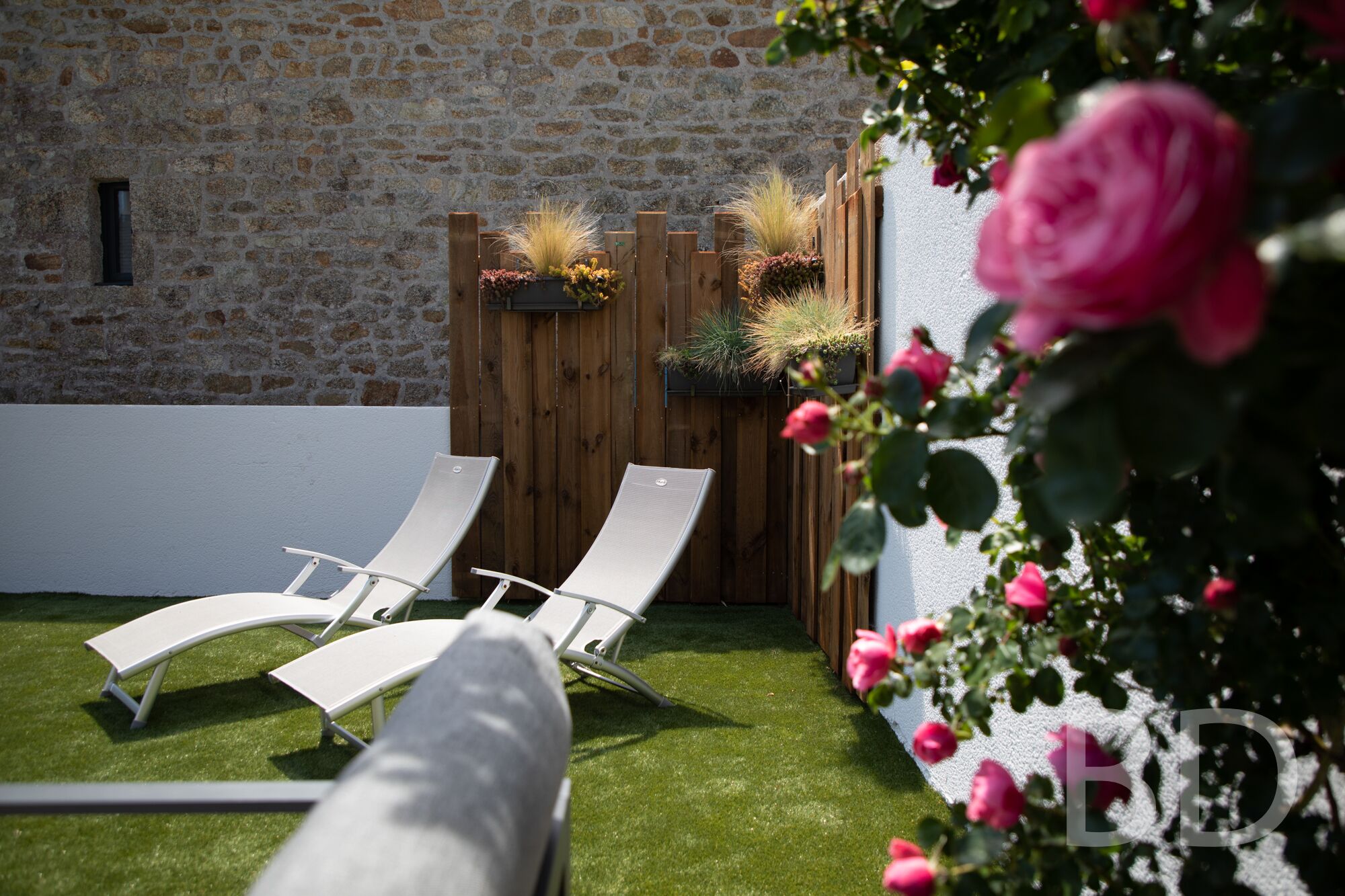 A patio with two white lounge chairs, a stone wall, and a wooden fence with plants.