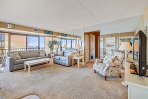 Ein geräumiges Wohnzimmer mit einem grauen Sofa, einem weißen Sessel und einem Couchtisch. Zwei Lampen stehen auf Beistelltischen, und ein großes Fenster bietet einen Blick ins Freie.