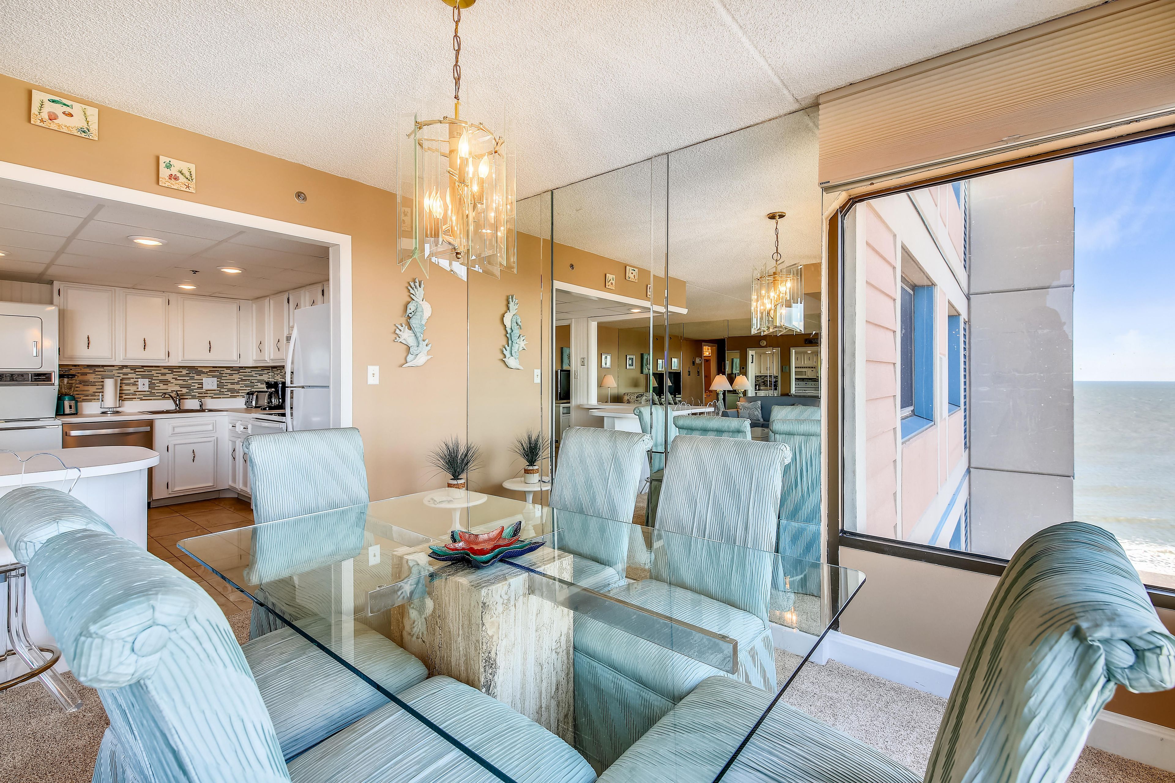 A modern interior with a glass dining table surrounded by striped chairs, a kitchen in the background, and a large window offering a view.