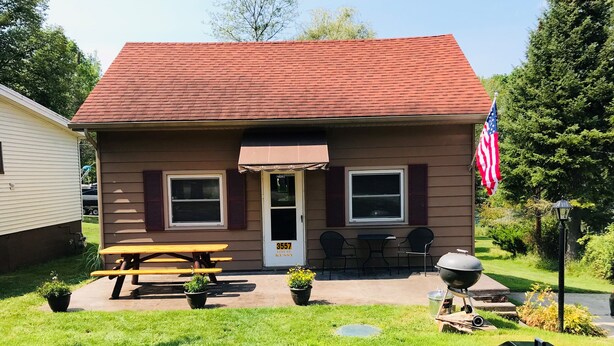 A small house with a red roof, a front door, two windows, a picnic table, and a grill. - Beautiful Year-Round  Home on the South Shore of Hubbard Lake (Spruce)