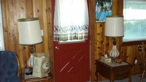 A room with a wooden paneled wall, a red door, two lamps, a small wooden table with a bag, a blue chair, and a patterned rug. - Beautiful Year-Round  Home on the South Shore of Hubbard Lake (Spruce)