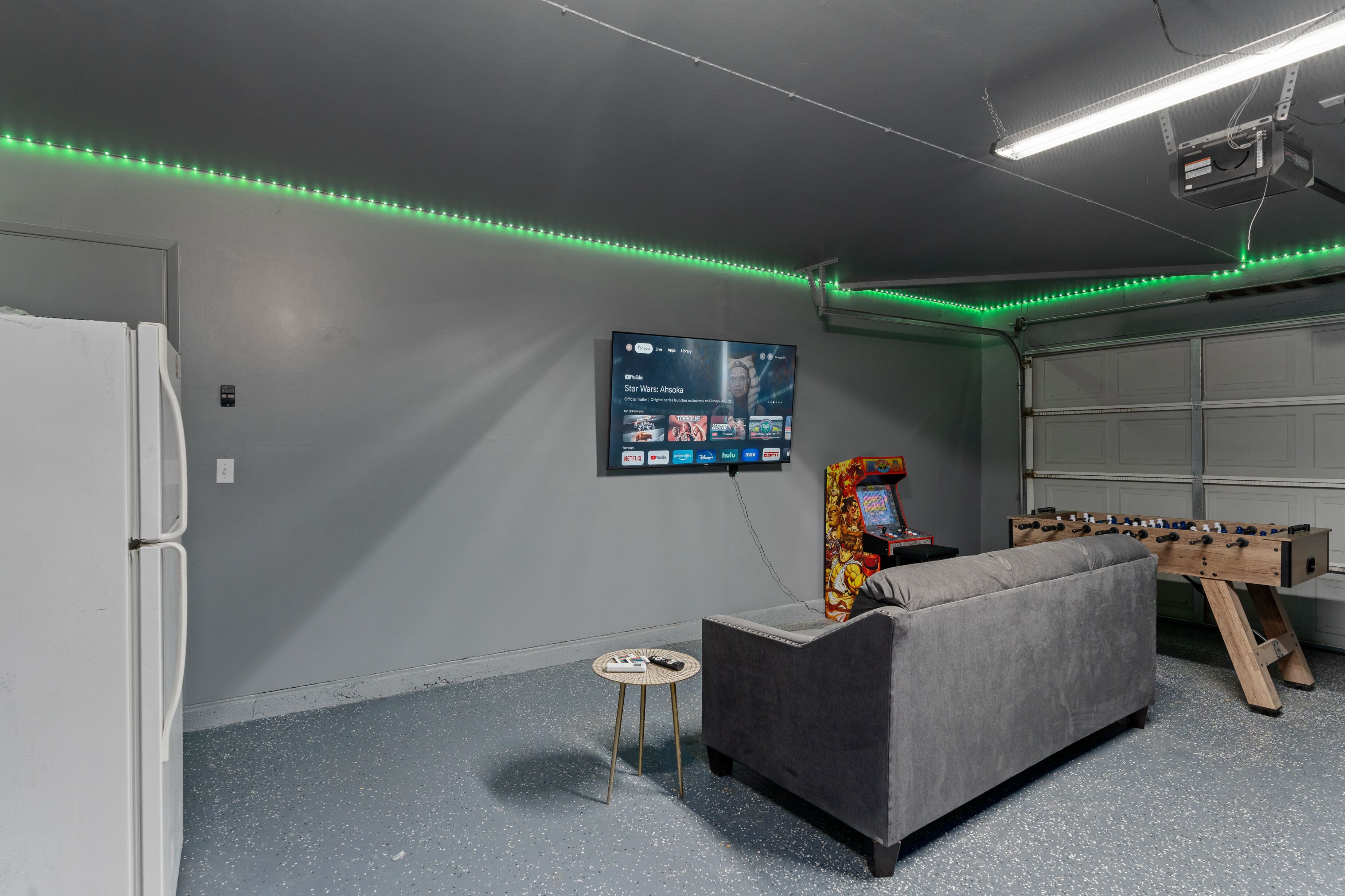 A room with a grey sofa, a foosball table, a TV mounted on the wall, and green LED strip lights along the ceiling.