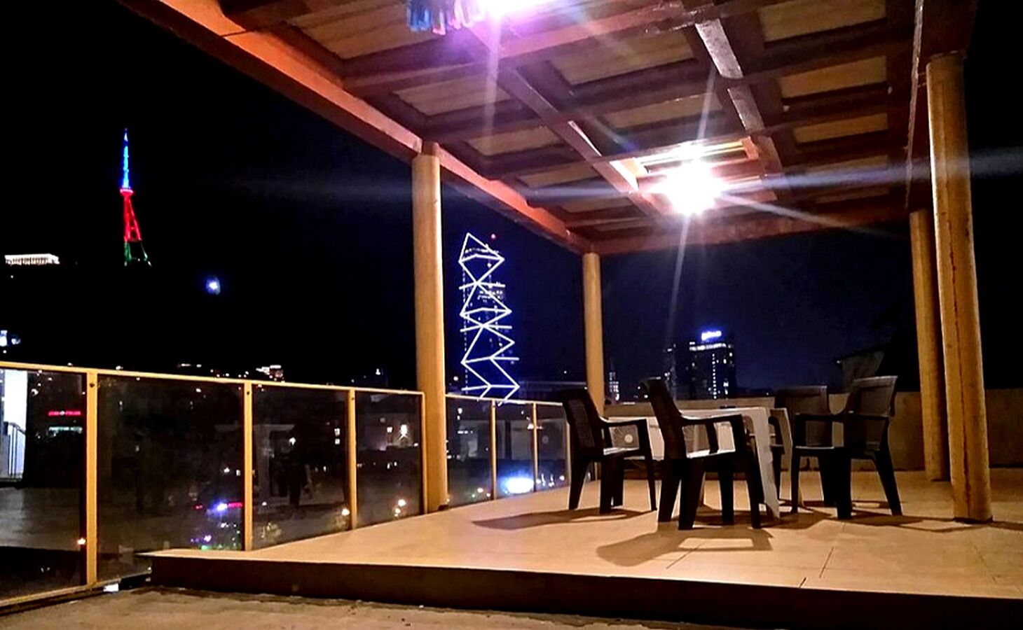 Un patio al aire libre con una mesa y sillas, barandilla de cristal y vista a un paisaje urbano nocturno.