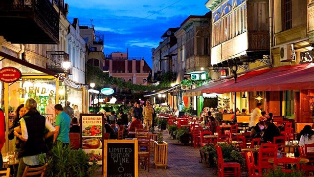 Une rue animée, avec des terrasses de cafés et de restaurants, une enseigne indiquant un restaurant de cuisine géorgienne et un panneau noir annonçant une dégustation de vin en édition limitée.