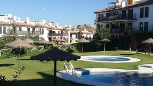 A swimming pool with a slide, surrounded by grass and thatched umbrellas, with buildings in the background.