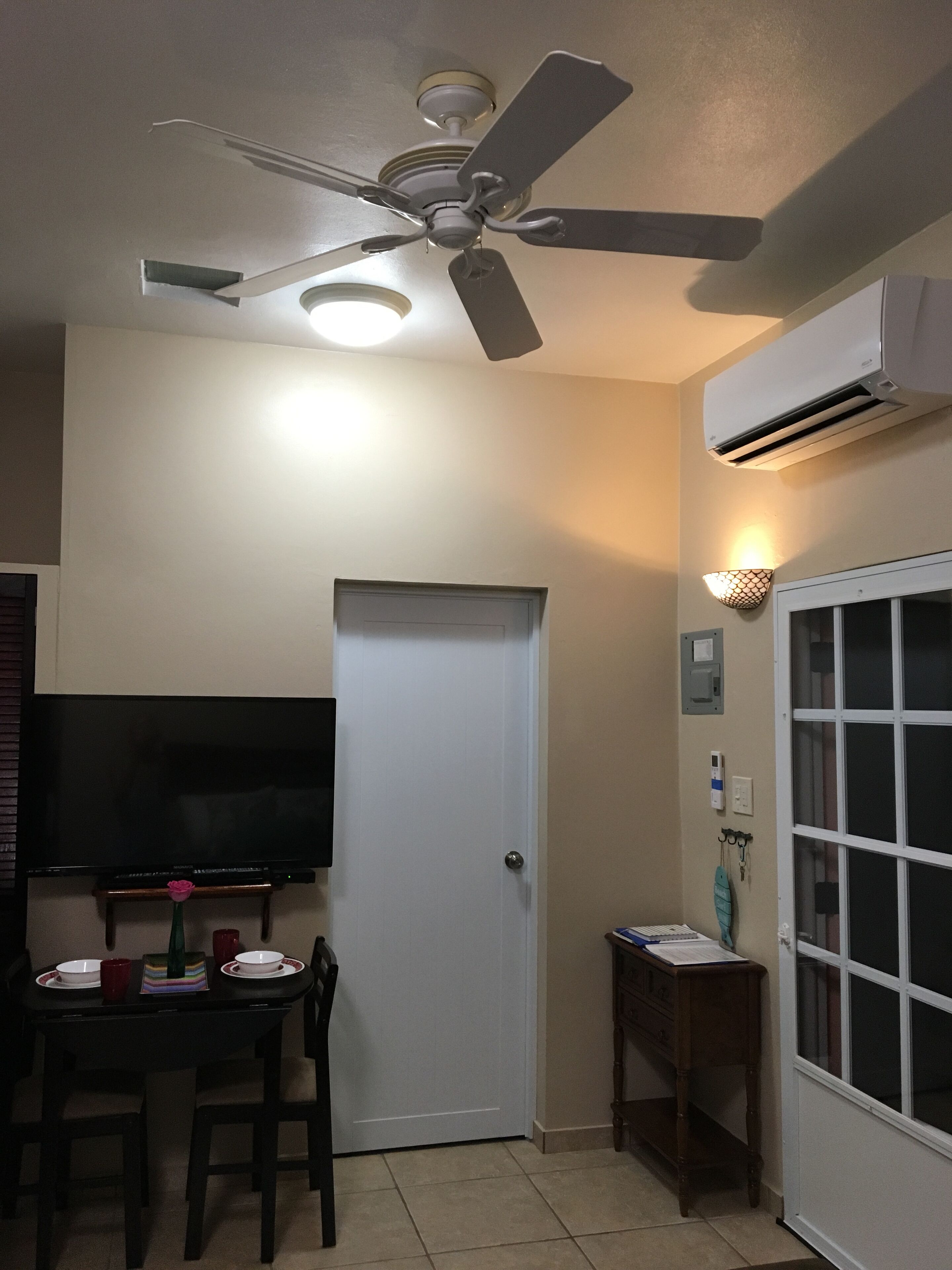 A room with a ceiling fan, a flat-screen TV mounted on the wall, a dining table with chairs, a white door, and an air conditioning unit.