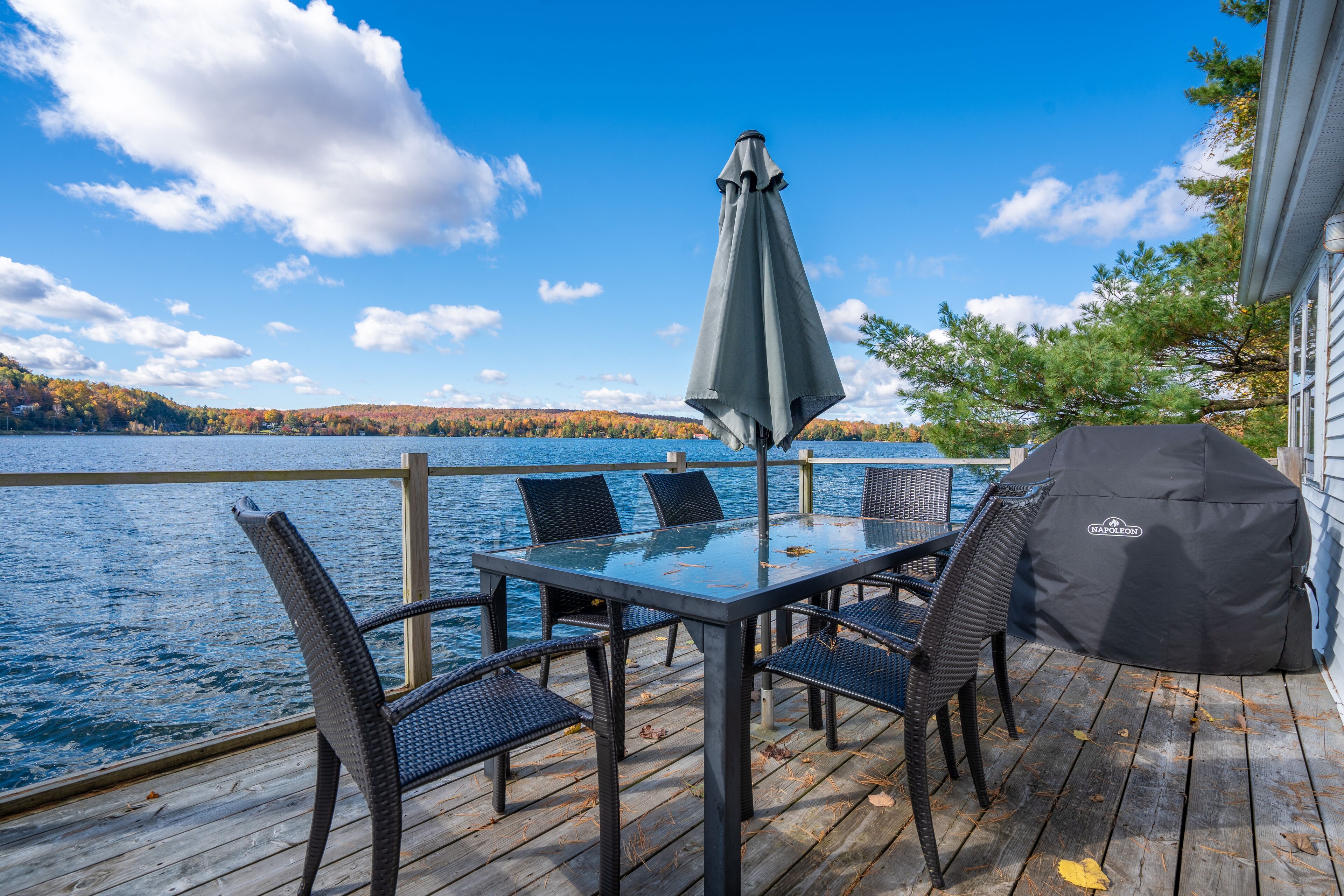 Un pontile in legno con tavolo e sedie in vetro, un barbecue e vista su un lago.