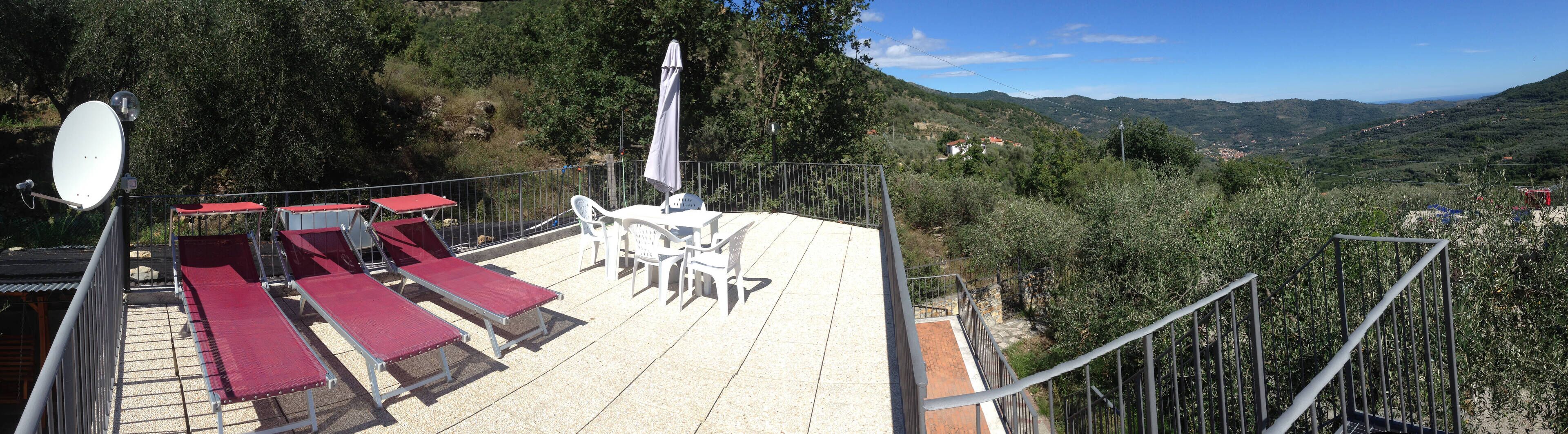A terrace with sun loungers and a table, surrounded by a metal railing, overlooking a mountainous landscape.